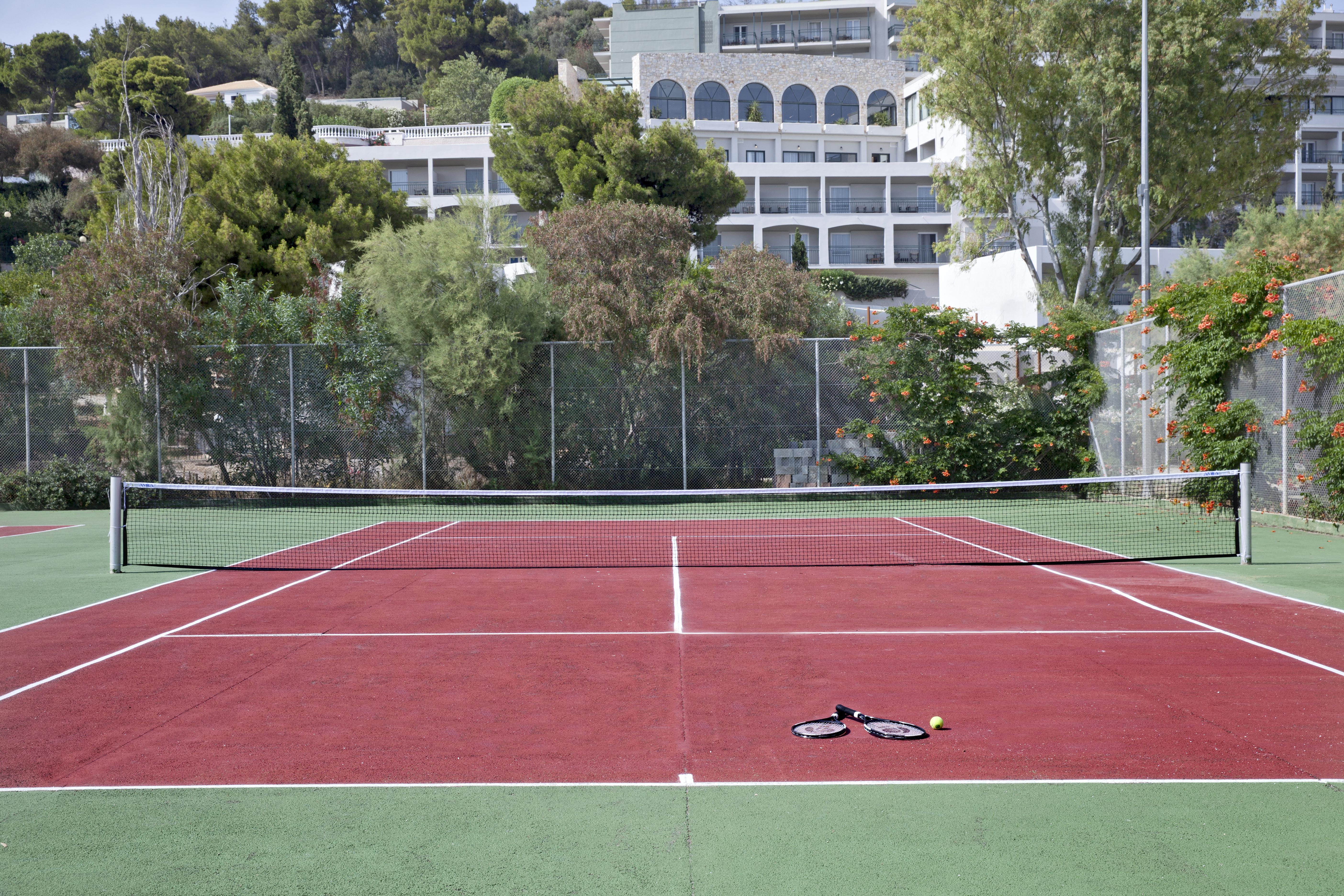 โดลเช บาย วินด์แฮม อัตติกา ริเวียรา Hotel วราโวนา ภายนอก รูปภาพ The photo shows a tennis court with red and green surfaces. The court is empty, with no players present. In the center, there are a tennis racket and a ball lying on the ground. In the background, there are trees and a building, suggesting the court 