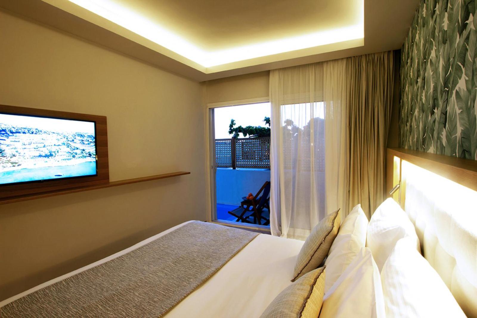 โดลเช บาย วินด์แฮม อัตติกา ริเวียรา Hotel วราโวนา ภายนอก รูปภาพ The photo shows a modern hotel room featuring a large bed with white linens and decorative pillows. There's a mounted flat-screen television on the wall opposite the bed. Natural light enters the room through sheer curtains, leading to a balcony or o
